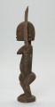 rzeźbiona figura - Ujęcie lewego boku. Drewniana, rzeźbiona postać kobiety. Jej lewa ręka podniesiona jest do góry.