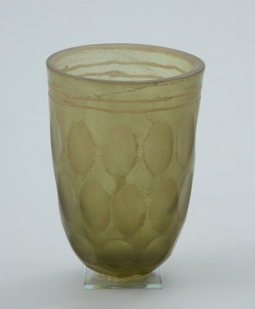 Pucharek szklany barwy jasnozielonej - Ujęcie z przodu. Pucharek szklany barwy jasnozielonej. Naczynie jest grubościenne zdobione czterema rzędami szlifowanych owali obiegających pucharek dookoła oraz dwoma żłobieniami pod krawędzią.