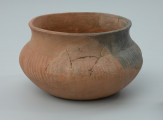 Naczynie gliniane (waza) - Ujęcie z przodu z lewej strony. Naczynie gliniane (waza) o przysadzistych proporcjach, barwy jasnobrunatnej. Brzusiec wazy jest w całości zdobiony dookolnie nieregularnymi skośnymi płytkimi bruzdami.