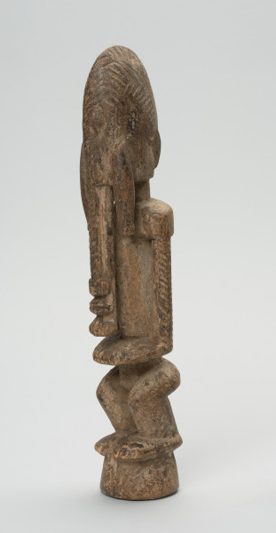 drewniana, rzeźbiona figura - Ujęcie z przodu, z lewej strony. Drewniana, rzeźbiona postać ludzka. Twarz schematycznie zaznaczona, zakończona bardzo długą brodą/podbródkiem. Po obu stronach brody dwa długie wąsy.