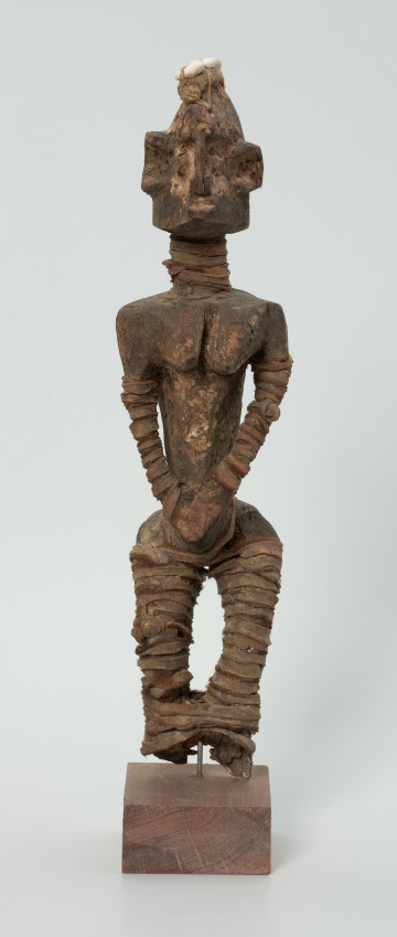 drewniana figura - Ujęcie z przodu. Drewniana, rzeźbiona postać kobiety, która wokół nóg i rąk ma obwiązane wąskie paski skóry. Do głowy przymocowany jest pasek bawełny z naszytymi muszelkami kauri.
