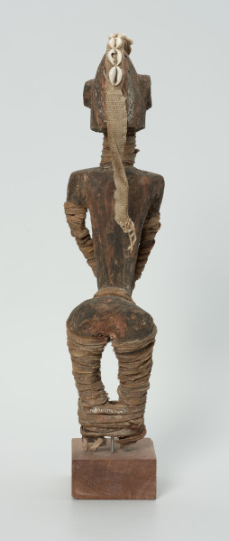drewniana figura - Ujęcie z tyłu. Drewniana, rzeźbiona postać kobiety, która wokół nóg i rąk ma obwiązane wąskie paski skóry. Do głowy przymocowany jest pasek bawełny z naszytymi muszelkami kauri.