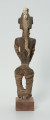 drewniana figura - Ujęcie z tyłu. Drewniana, rzeźbiona postać kobiety, która wokół nóg i rąk ma obwiązane wąskie paski skóry. Do głowy przymocowany jest pasek bawełny z naszytymi muszelkami kauri.