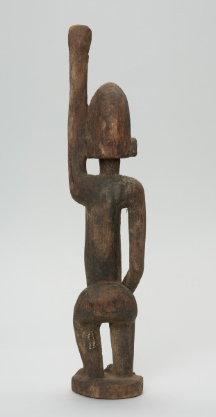 rzeźbiona figura - Ujęcie z tyłu. Drewniana, rzeźbiona postać kobiety. Jej lewa ręka podniesiona jest do góry.