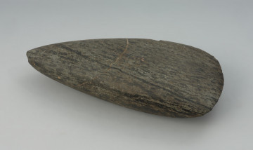 Siekiera kamienna - Ujęcie z góry; Siekiera kamienna o owalnym przekroju poprzecznym, ze spiczastym obuchem oraz z nieco asymetrycznym, łukowatym ostrzem. Powierzchnia jest silnie zeszlifowana, równa i gładka. Wykonana jest z surowca skalnego o zielonkawej barwie z czarnymi pasmami