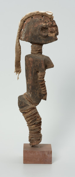 drewniana figura - Ujęcie prawego boku. Drewniana, rzeźbiona postać kobiety, która wokół nóg i rąk ma obwiązane wąskie paski skóry. Do głowy przymocowany jest pasek bawełny z naszytymi muszelkami kauri.