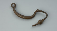 Kabłąk zapinki z brązu - Ujęcie z góry z przodu. Zapinka z brązu; zachował się tylko kabłąk jednoczęściowy, przechodzący w odgiętą w górę nóżkę, zachowany jest jeden zwój sprężynki.