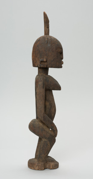 rzeźbiona figura - Ujęcie prawego boku. Drewniana, rzeźbiona postać kobiety. Jej lewa ręka podniesiona jest do góry.