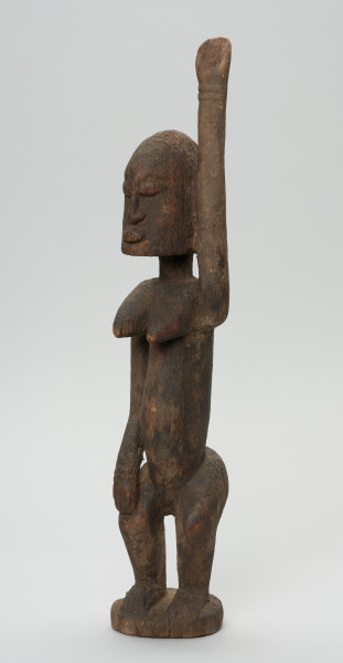 rzeźbiona figura - Ujęcie z przodu, z lewej strony. Drewniana, rzeźbiona postać kobiety. Jej lewa ręka podniesiona jest do góry.