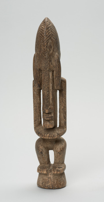 drewniana, rzeźbiona figura - Ujęcie z przodu. Drewniana, rzeźbiona postać ludzka. Twarz schematycznie zaznaczona, zakończona bardzo długą brodą/podbródkiem. Po obu stronach brody dwa długie wąsy.