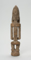 drewniana, rzeźbiona figura - Ujęcie z tyłu. Drewniana, rzeźbiona postać ludzka. Twarz schematycznie zaznaczona, zakończona bardzo długą brodą/podbródkiem. Po obu stronach brody dwa długie wąsy.