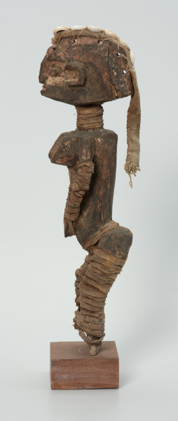 drewniana figura - Ujęcie lewego boku. Drewniana, rzeźbiona postać kobiety, która wokół nóg i rąk ma obwiązane wąskie paski skóry. Do głowy przymocowany jest pasek bawełny z naszytymi muszelkami kauri.