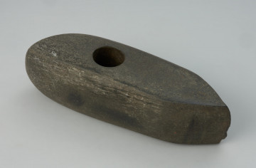 Topór kamienny - Ujęcie z przodu; Asymetryczne narzędzie ze skośnym obuchem i otworem przewierconym równolegle do kierunku ostrza. Otwór do osadzenia trzonka umieszczony bliżej części tylnej, jest wywiercony jednostronnie. W partii środkowej, opodal otworu, jest widoczne zaczątkowe wiercenie wykonywane okrągłym, pustym narzędziem, prawdopodobnie kością. Powierzchnia narzędzia jest gładzona w nierównym stopniu, przy otworze zauważalne są zagłębienia po odbiciach kształtujących ogólną formę narzędzia. W wielu miejscach zaobserwować można grupy równoległych rys, które są śladami po zdzieraniu i gładzeniu