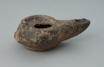 Rzymska lampka oliwna z gliny z urwanym uchem - Ujęcie z boku. Rzymska lampka oliwna z gliny z urwanym uchem.