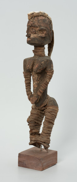 drewniana figura - Ujęcie z przodu, z lewej strony. Drewniana, rzeźbiona postać kobiety, która wokół nóg i rąk ma obwiązane wąskie paski skóry. Do głowy przymocowany jest pasek bawełny z naszytymi muszelkami kauri.