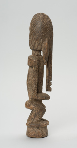 drewniana, rzeźbiona figura - Ujęcie prawego boku. Drewniana, rzeźbiona postać ludzka. Twarz schematycznie zaznaczona, zakończona bardzo długą brodą/podbródkiem. Po obu stronach brody dwa długie wąsy.