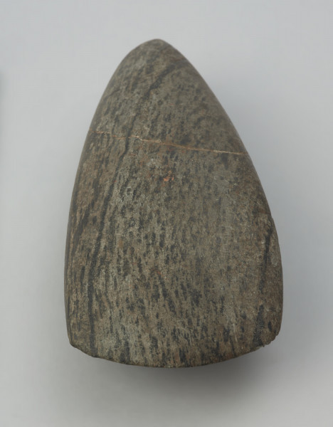 Siekiera kamienna - Ujęcie w pionie ostrzem do góry; Siekiera kamienna o owalnym przekroju poprzecznym, ze spiczastym obuchem oraz z nieco asymetrycznym, łukowatym ostrzem. Powierzchnia jest silnie zeszlifowana, równa i gładka. Wykonana jest z surowca skalnego o zielonkawej barwie z czarnymi pasmami