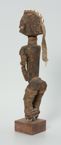 drewniana figura - Ujęcie z tyłu, z lewej strony. Drewniana, rzeźbiona postać kobiety, która wokół nóg i rąk ma obwiązane wąskie paski skóry. Do głowy przymocowany jest pasek bawełny z naszytymi muszelkami kauri.