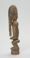 drewniana, rzeźbiona figura - Ujęcie lewego boku. Drewniana, rzeźbiona postać ludzka. Twarz schematycznie zaznaczona, zakończona bardzo długą brodą/podbródkiem. Po obu stronach brody dwa długie wąsy.