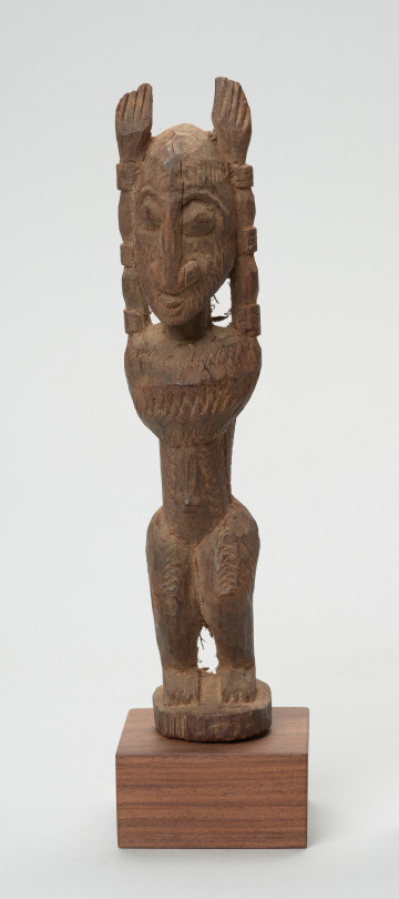 rzeźba - Ujęcie z przodu. Figura mężczyzny w pozycji stojącej (na ugiętych w kolanach nogach) z rękoma uniesionymi do góry. Cała powierzchnia rzeźby pokryta jest delikatnym rytem, sugerującym skaryfikację.