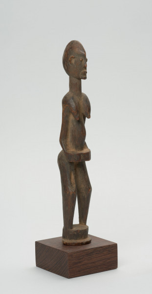 rzeźba - Ujęcie ze skosu z lewej; Figura - postać kobiety w pozycji stojącej. Głowa mała, podłużna, ręce, piersi, brzuch lekko wydobyte z bryły figury. Nogi bez stóp, przechodzą od razu w okrągły cokolik.