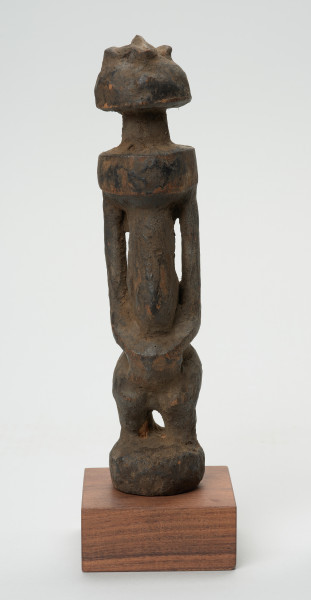 rzeźba - Ujęcie z przodu. Rzeźba w drewnie w formie uproszczonej. Przedstawia postać męską o lekko zarysowanej głowie, szczupłym, długim tułowiu, krótkie nogi ugięte, ręce oparte na kolanach.
