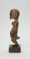 rzeźba - Ujęcie prawej strony rzeźby. Drewniana rzeźba postaci mężczyzny w pozycji siedzącej. Głowa duża z zaznaczonym długim cienkim nosem, strzałkowatym, długie ręce oparte na kolanach, krótkie nogi.