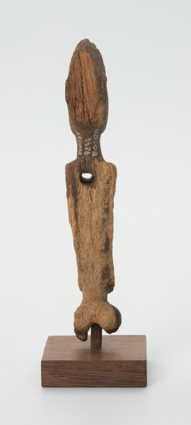 Drewniana figurka kobiety z otworem w lewej piersi - Ujęcie z tyłu. Drewniana figurka postaci kobiecej, na której widoczne są piersi.  Ręce lekko zaznaczone, ułożone wzdłuż tułowia. Brak nóg. Na wysokości lewej piersi otwór.