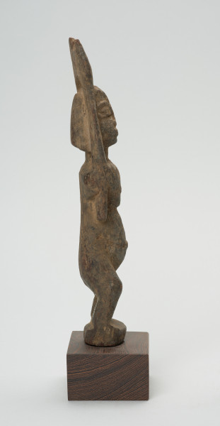 rzeźba - Ujęcie lewej strony rzeźby. Drewniana rzeźba postaci kobiety. Długie ręce wzniesione do góry, głowa podłużna, wyraźnie zaznaczone duże oczy, zarysowany wypukły brzuch, krótkie, lekko skrzywione nogi.