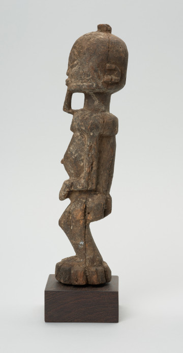 rzeźba - Ujęcie lewego boku. Figura przedstawiająca mężczyznę stojącego na nogach ugiętych w kolanach. Głowa duża, zaokrąglona, oczy duże, ze źrenicami, wyraźnie zaznaczona bródka.