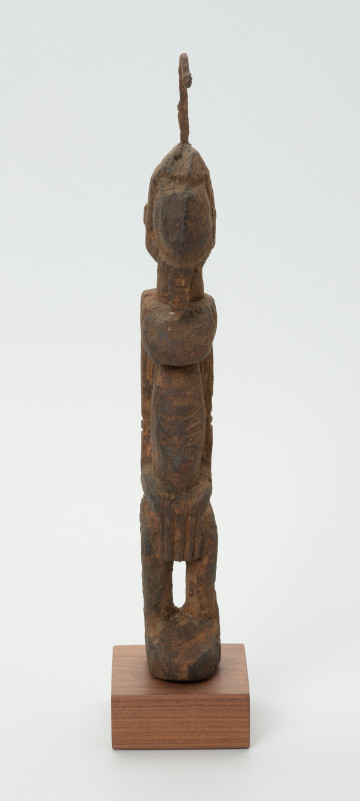 rzeźba - Ujęcie z przodu. Drewniana rzeźba postaci męskiej. Wąska podłużna twarz, na głowie haczykowata ozdoba. Tułów długi, nogi zgięte w kolanach, ręce oparte na kolanach.