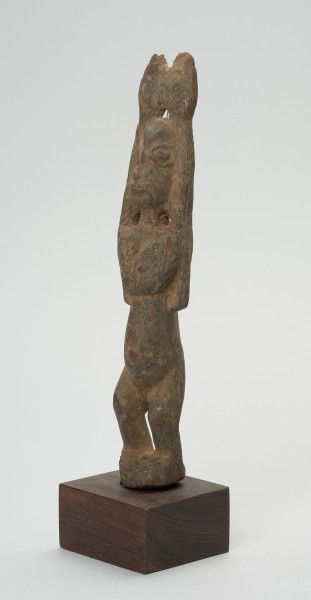rzeźba - Ujęcie przodu skosem w lewą stronę. Drewniana rzeźba postaci kobiety. Długie ręce wzniesione do góry, głowa podłużna, wyraźnie zaznaczone duże oczy, zarysowany wypukły brzuch, krótkie, lekko skrzywione nogi.