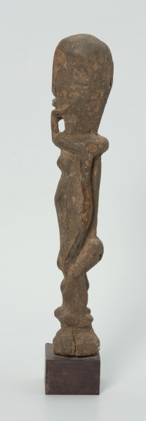Drewniana figurka androgyniczna - Ujęcie z lewej strony; drewniana figurka przedstawiająca postać z brodą i piersiami. Statyczna, zgeometryzowana. Ręce wyodrębnione z tułowia.