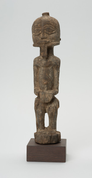 rzeźba - Ujęcie z przodu. Figura przedstawiająca mężczyznę stojącego na nogach ugiętych w kolanach. Głowa duża, zaokrąglona, oczy duże, ze źrenicami, wyraźnie zaznaczona bródka.