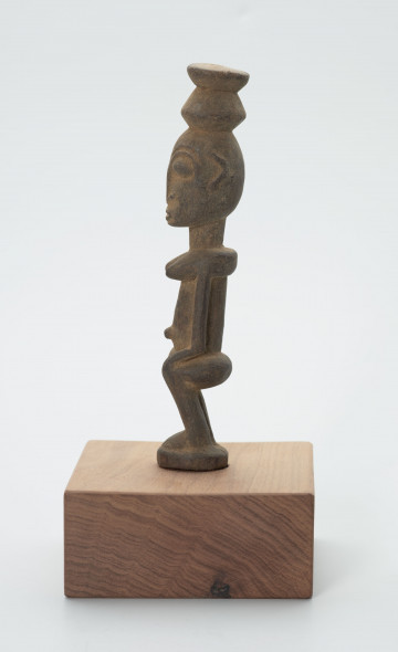 rzeźba - Ujęcie z prawego boku; Figura - postać kobiety. Postać w pozycji siedzącej. Ręce oparte na kolanach. Na głowie naczynie. Bardzo uwydatniony pępek. Znacząco płaska twarz.