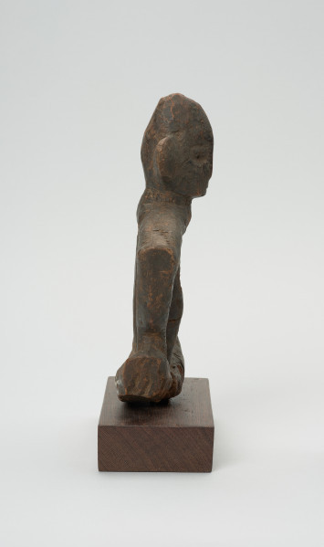 rzeźba - Ujęcie z lewej strony. Figurka przedstawiająca postać ludzką. Ręce i nogi w przysiadzie wąskie, połączone ze sobą w formę kwadratu. Głowa spiczasta z odstającymi szerokimi uszami. Tułów cienki, prosty.