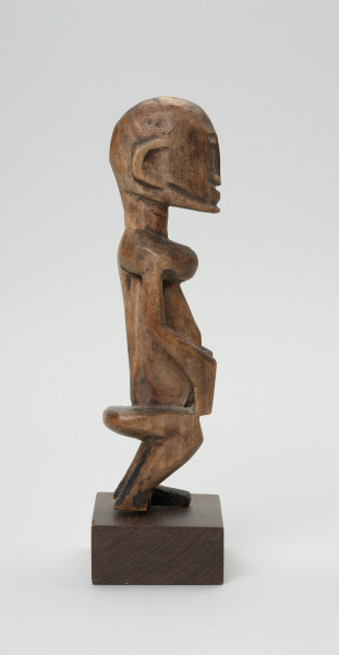 rzeźba - Ujęcie lewej strony rzeźby. Drewniana rzeźba postaci mężczyzny w pozycji siedzącej. Głowa duża z zaznaczonym długim cienkim nosem, strzałkowatym, długie ręce oparte na kolanach, krótkie nogi.