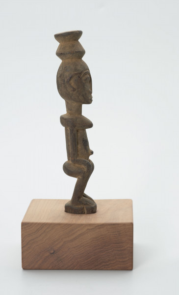 rzeźba - Ujęcie z lewego boku; Figura - postać kobiety. Kobieta w pozycji siedzącej. Ręce spoczywaja na kolanach. Na głowie naczynie. Bardzo wypukły pępek. Twarz płaska.