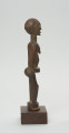 rzeźba - Ujęcie z lewego boku; Figura - postać kobiety w pozycji stojącej. Głowa mała, podłużna, ręce, piersi, brzuch lekko wydobyte z bryły figury. Nogi bez stóp, przechodzą od razu w okrągły cokolik.
