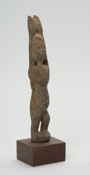 rzeźba - Ujęcie przodu skosem w prawą stronę. Drewniana rzeźba postaci kobiety. Długie ręce wzniesione do góry, głowa podłużna, wyraźnie zaznaczone duże oczy, zarysowany wypukły brzuch, krótkie, lekko skrzywione nogi.