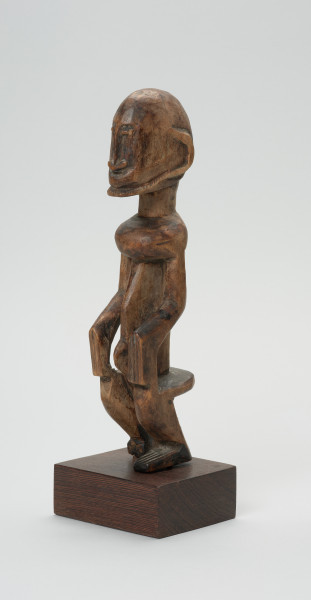 rzeźba - Ujęcie przodu skosem w lewą stronę. Drewniana rzeźba postaci mężczyzny w pozycji siedzącej. Głowa duża z zaznaczonym długim cienkim nosem, strzałkowatym, długie ręce oparte na kolanach, krótkie nogi.