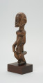 rzeźba - Ujęcie przodu skosem w lewą stronę. Drewniana rzeźba postaci mężczyzny w pozycji siedzącej. Głowa duża z zaznaczonym długim cienkim nosem, strzałkowatym, długie ręce oparte na kolanach, krótkie nogi.