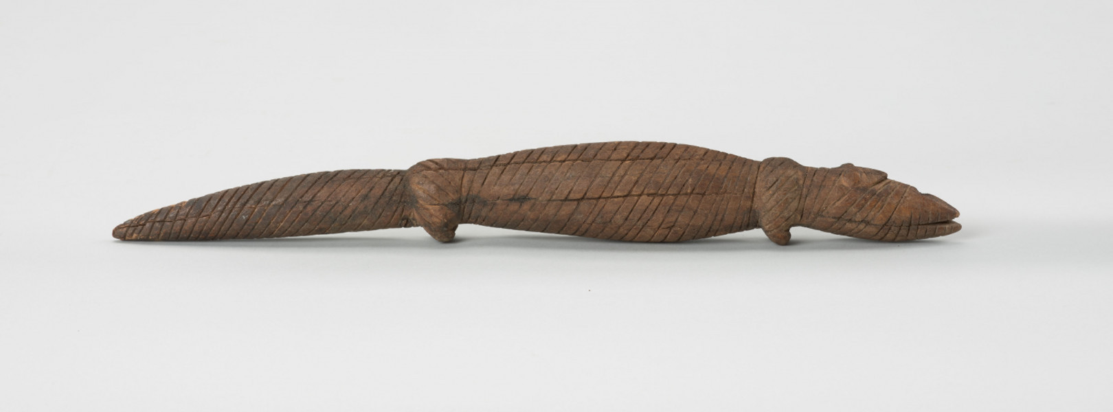 Drewniana figurka krokodyla - Ujęcie z prawej strony; Drewniana figurka krokodyla, na całej długości pokryta drobnymi nacięciami. Zaznaczona długa głowa, długi tułów oraz gruby ogon, zakończony spiczaście i spuszczony do dołu. Zaznaczone proste, krótkie kończyny przednie i tylne.