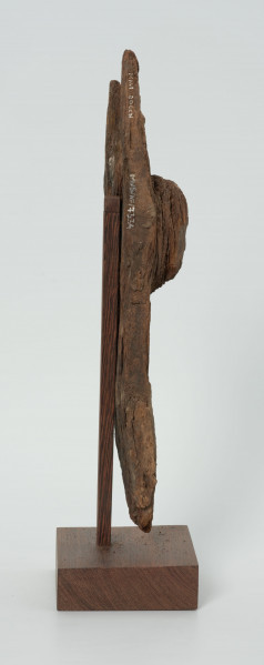 Drewniana figurka na rozwidlonej u góry desce - Ujęcie z prawej strony; na płaskiej, rozwidlonej na szczycie desce wyrzeźbiono okrągłą głowę z delikatnie zaznaczonymi oczami, nosem z małymi ustami. Figura posiada liczne spękania.