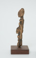 rzeźba - Ujęcie prawej strony rzeźby. Drewniana rzeźba dwóch postaci. Schematycznie zarysowana postać z podłużną głową, szczupłym tułowiem i krótkimi nogami. Większa postać niesie drugą mniejszą z przodu.