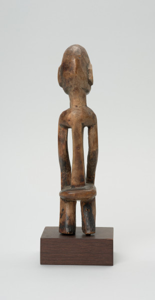 rzeźba - Ujęcie z tyłu. Drewniana rzeźba postaci mężczyzny w pozycji siedzącej. Głowa duża z zaznaczonym długim cienkim nosem, strzałkowatym, długie ręce oparte na kolanach, krótkie nogi.