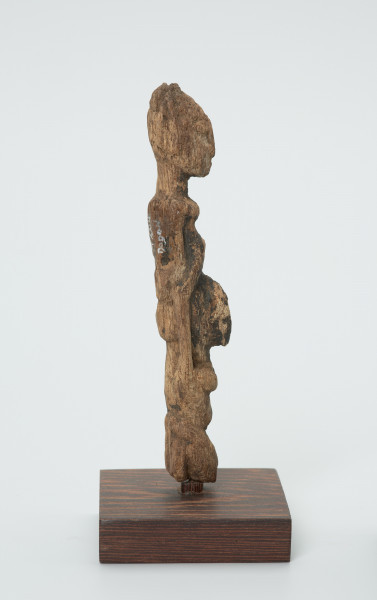 rzeźba - Ujęcie lewej strony rzeźby. Drewniana rzeźba dwóch postaci. Schematycznie zarysowana postać z podłużną głową, szczupłym tułowiem i krótkimi nogami. Większa postać niesie drugą mniejszą z przodu.