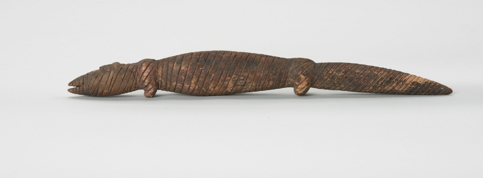 Drewniana figurka krokodyla - Ujęcie z lewej strony; Drewniana figurka krokodyla, na całej długości pokryta drobnymi nacięciami. Zaznaczona długa głowa, długi tułów oraz gruby ogon, zakończony spiczaście i spuszczony do dołu. Zaznaczone proste, krótkie kończyny przednie i tylne.