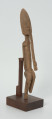 drewniana, rzeźbiona figura - Ujęcie z przodu, z prawej strony. Drewniana, rzeźbiona figura smukłej postaci znajdującej się w pozycji siedzącej. Długa broda ciągnąca się aż do kolan. Prawa dłoń spoczywa na kolanie, lewa trzyma brodę.