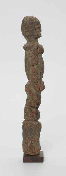 Drewniana figurka mężczyzny z twarzą zwróconą w prawo - Ujęcie z lewego boku; Lewy profil drewnianej figurki człowieka. Lewa ręka płaskorzeźbiona wzdłuż tułowia. Noga przechodzi w długi cokolik.